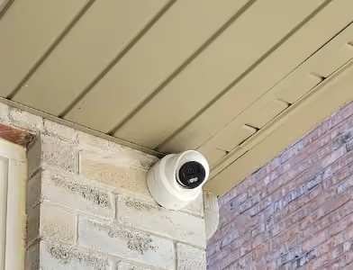 White outdoor security camera mounted beneath a porch soffit beside a brick wall.