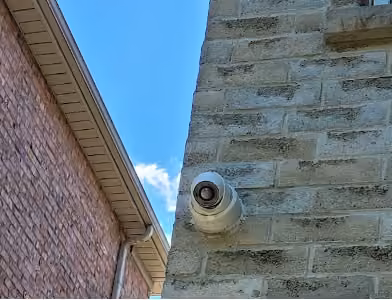 Security camera mounted on a light brick wall overlooking the passage between two houses.