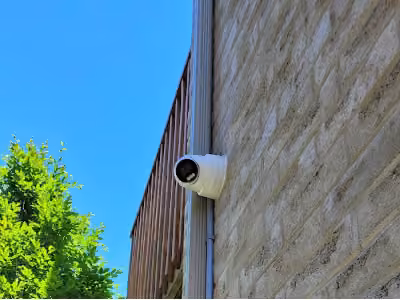 White outdoor security camera on an exterior brick wall beside a downspout.