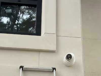 White security camera beneath a window on a light-coloured exterior wall.
